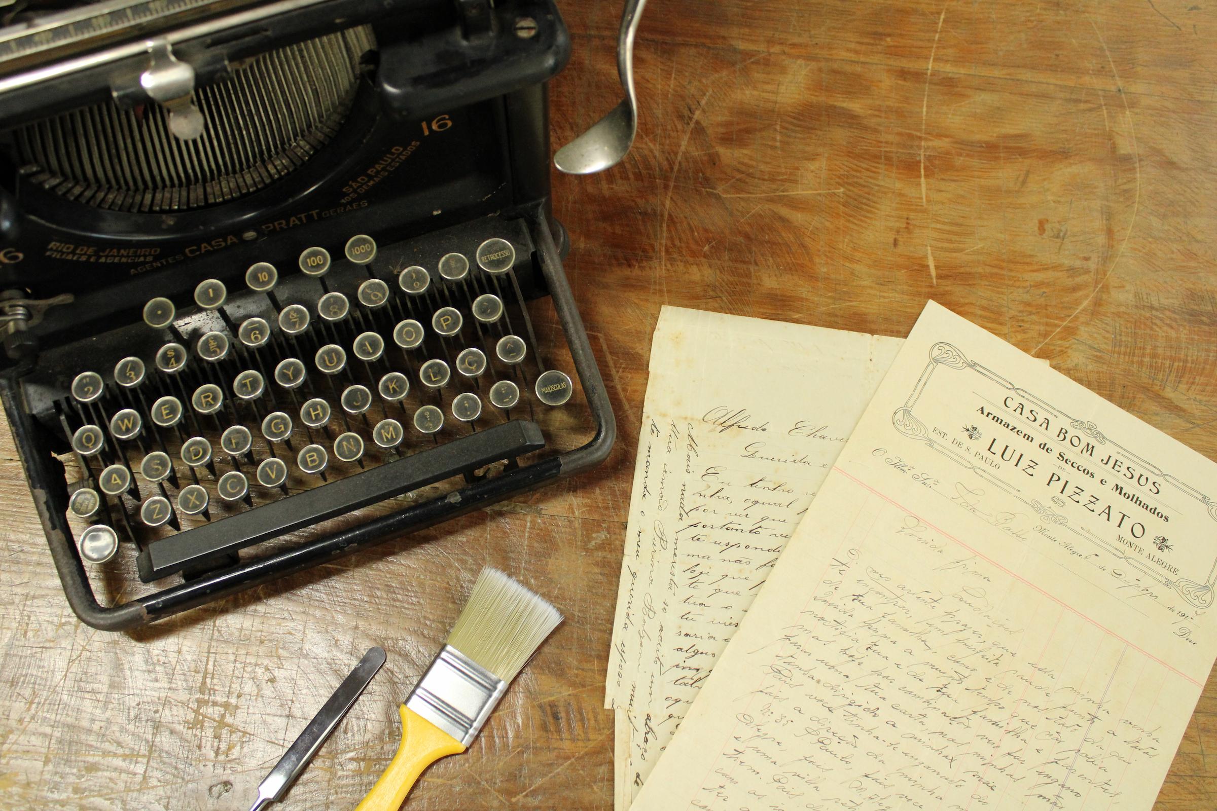 Foto de uma máquina de escrever antiga ao lado de alguns documentos antigos sobre uma mesa.