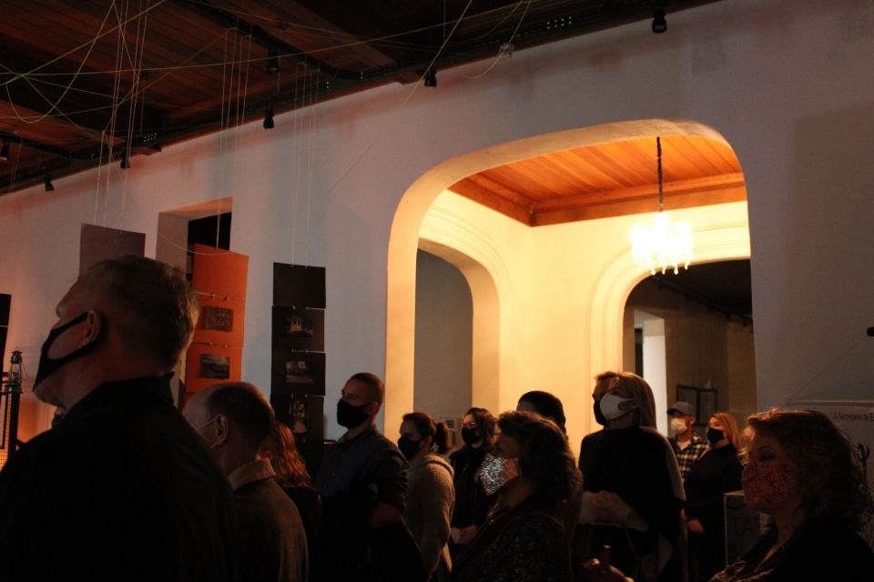 Fotografia de pessoas circulando em espaço de exposição. Em destaque, projeção de vídeo com entrevista na parede.