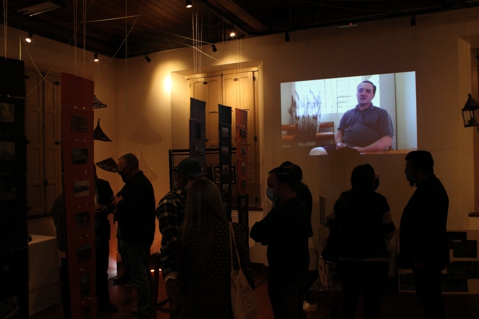 Fotografia de pessoas circulando em espaço de exposição. Em destaque, projeção de vídeo com entrevista na parede.