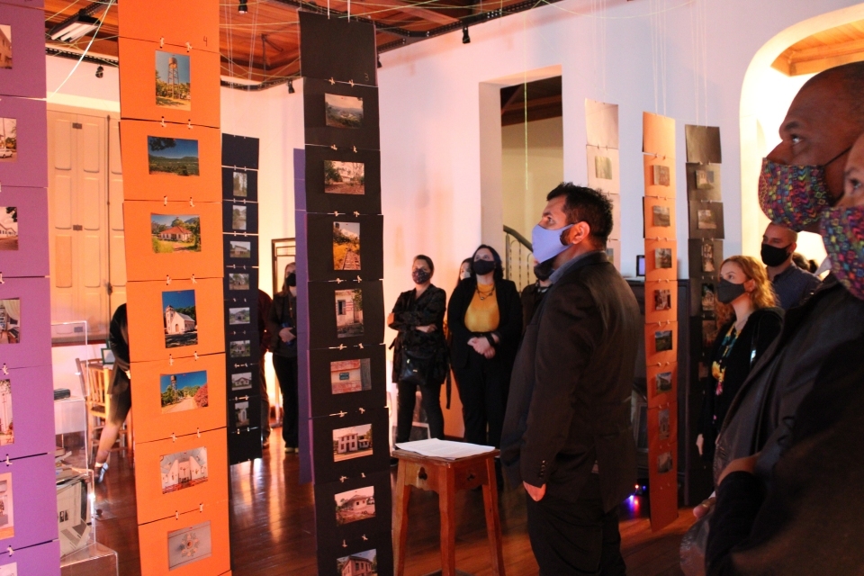 Fotografia de pessoas circulando em espaço de exposição. Em destaque, projeção de vídeo com entrevista na parede.