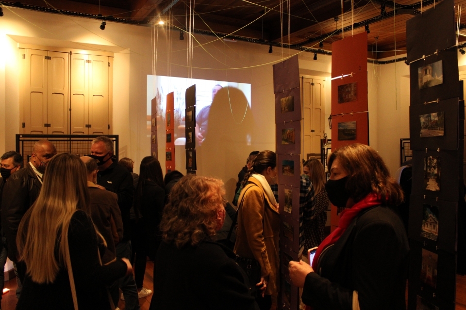 Fotografia de pessoas circulando em espaço de exposição. Em destaque, projeção de vídeo com entrevista na parede.