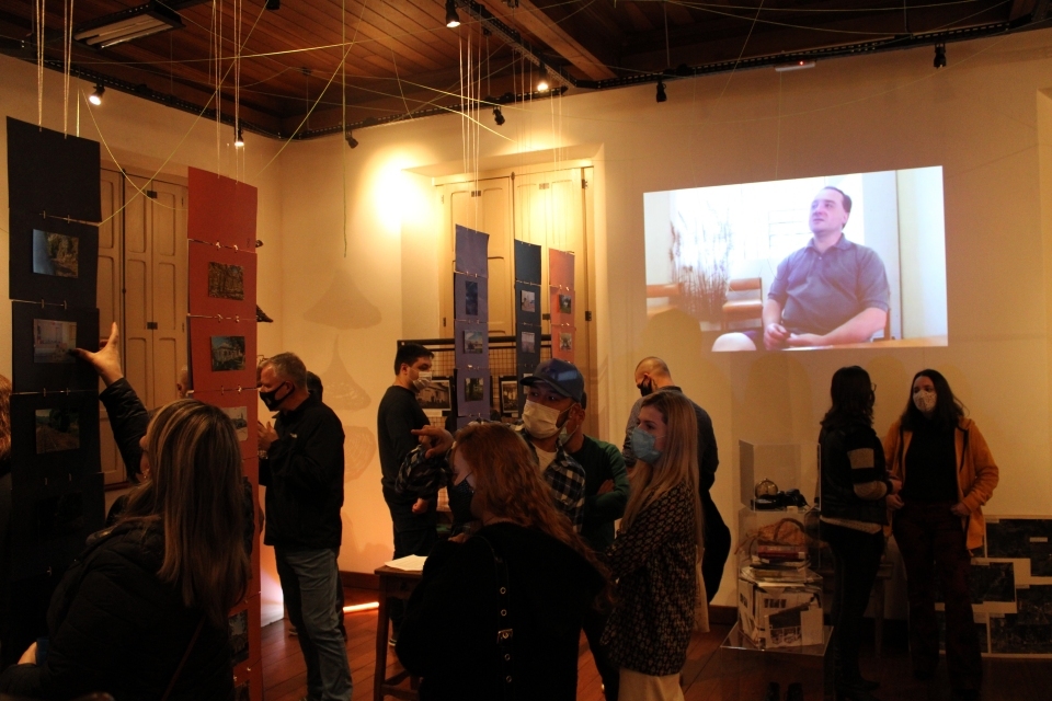 Fotografia de pessoas circulando em espaço de exposição. Em destaque, projeção de vídeo com entrevista na parede.