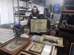 Fotografia de mulher segurando quadro. Em sua frente, diversos quadros e pinturas organizados em uma mesa de madeira.